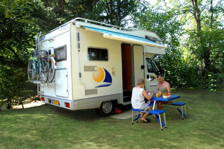 Emplacement camping-car Camping-car et ses vélos sur un emplacement ombragé, un couple déjeunant à une table pliante sur l'herbe